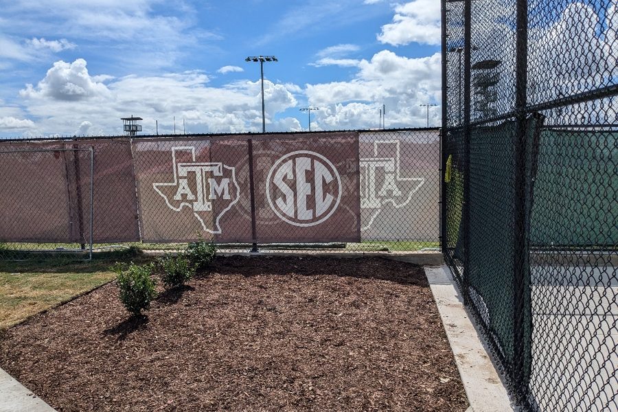 Black chain link fence with Texas A&M branded windscreen beside a mulched bed